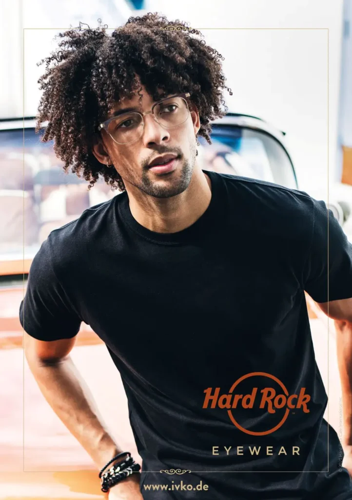 Ein Mann mit lockigen Haaren in einem schwarzen Shirt trägt eine helle Brille und lehnt an der Motorhaube eines Autos.