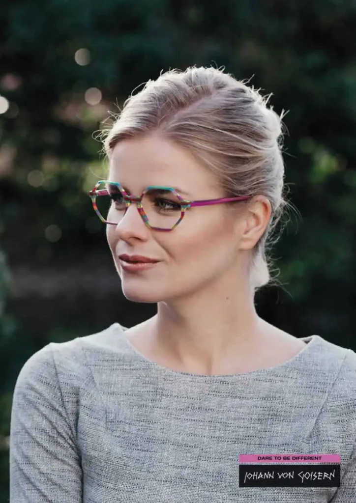 Eine blonde Frau trägt eine bunte Kunststoffbrille und einen grauen Pullover.