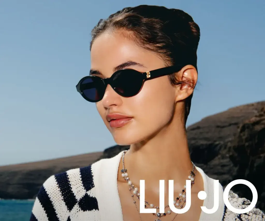 Eine dunkelhaarige Frau steht vor einer Bucht und trägt eine dunkle Sonnenbrille.