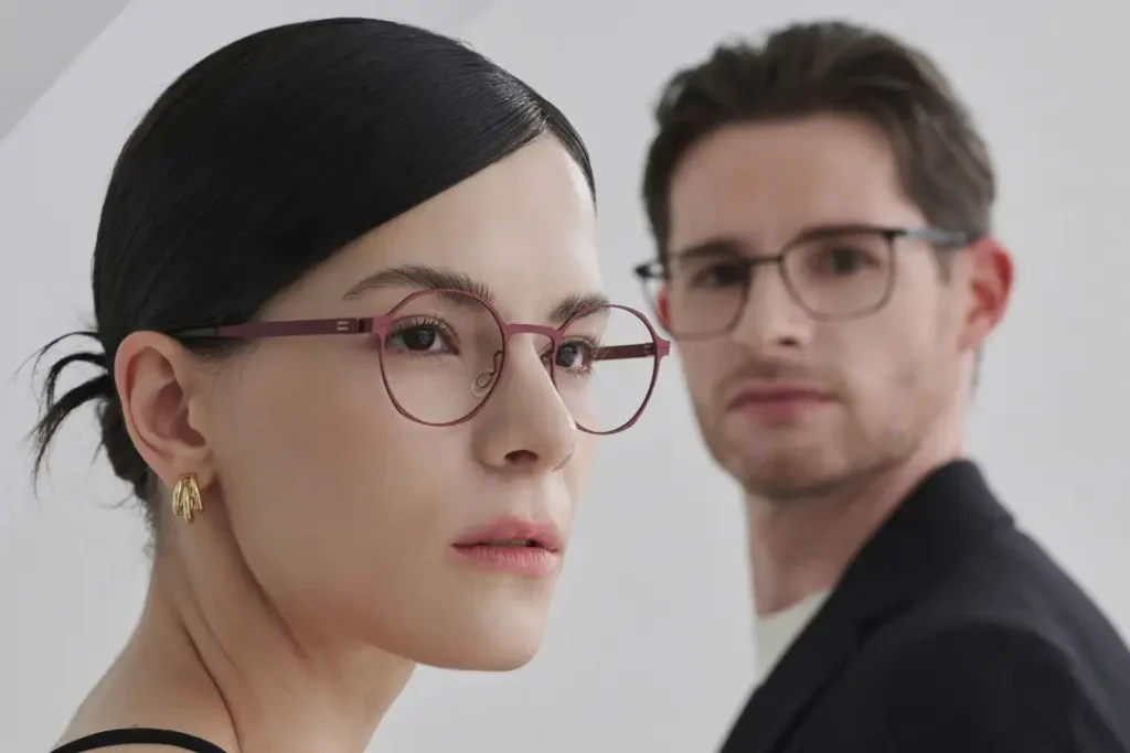 Ein dunkelhaariger Mann mit Brille und Anzug steht im Hintergrund von einer dunkelhaarigen Frau mit einer roten Brille.
