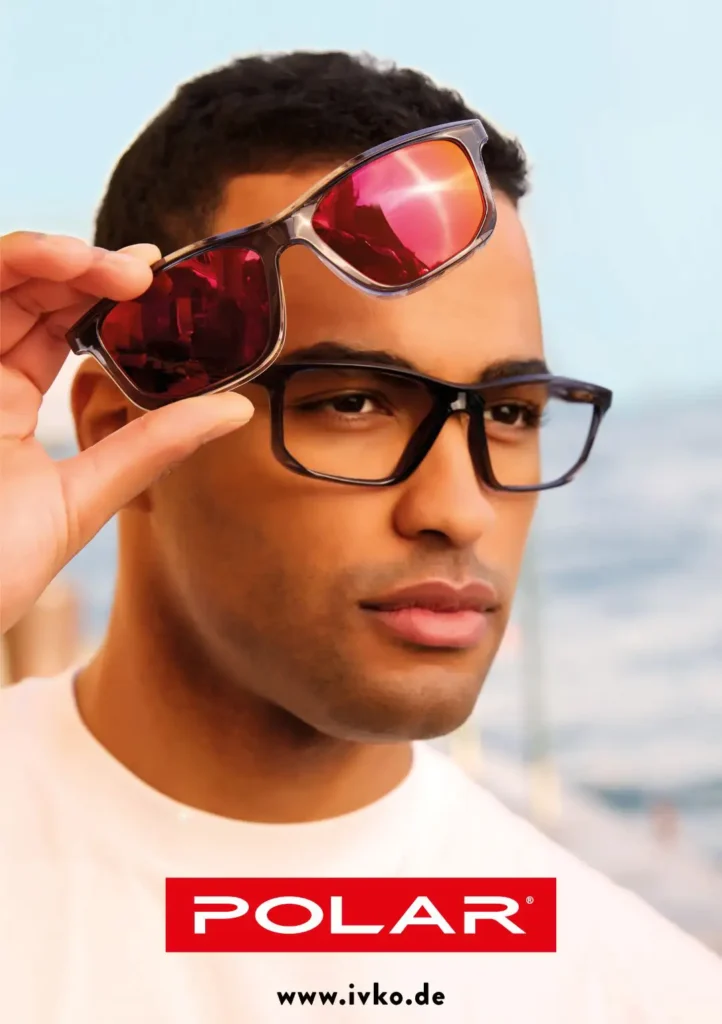 Dunkelhäutiger Mann mit kurzen schwarzen Haaren und einer schwarzen Kunststoffbrille mit einem verspiegelten Sonnenbrillenclip in der rechten Hand