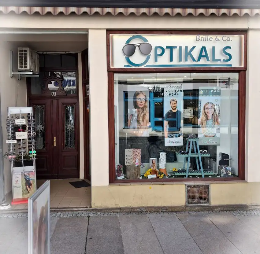 Schaufenster in dem Banner mit Brillenträgern hängen und Brillen ausgestellt sind. Darüber ist der leuchtende Schriftzug Optikals zu lesen. Im Eingangsbereich steht ein Ständer mit Sonnenbrillen.